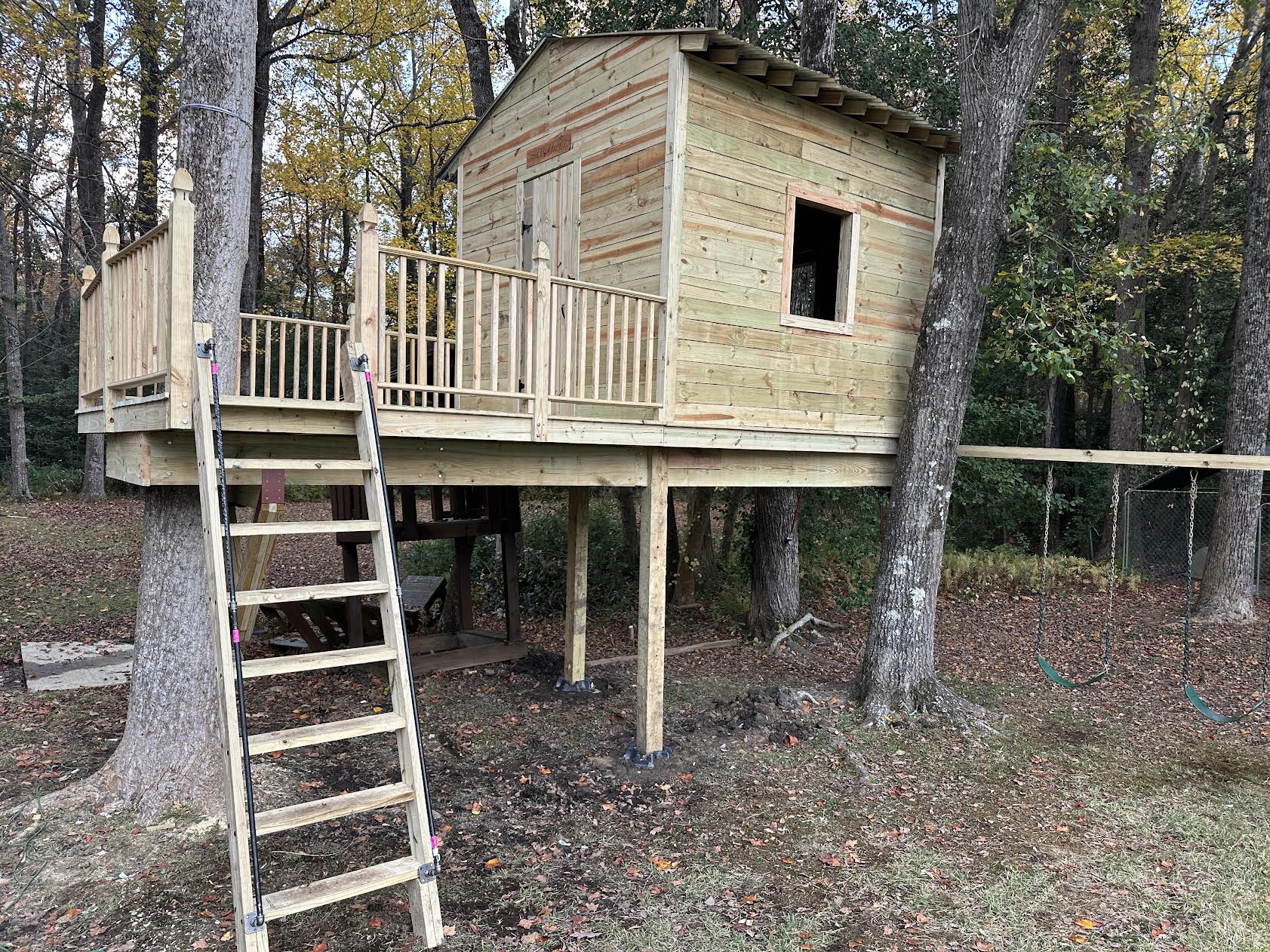 Finished cabin treehouse on posts between two trees, with a deck, railing, ladder and a swing set alongside