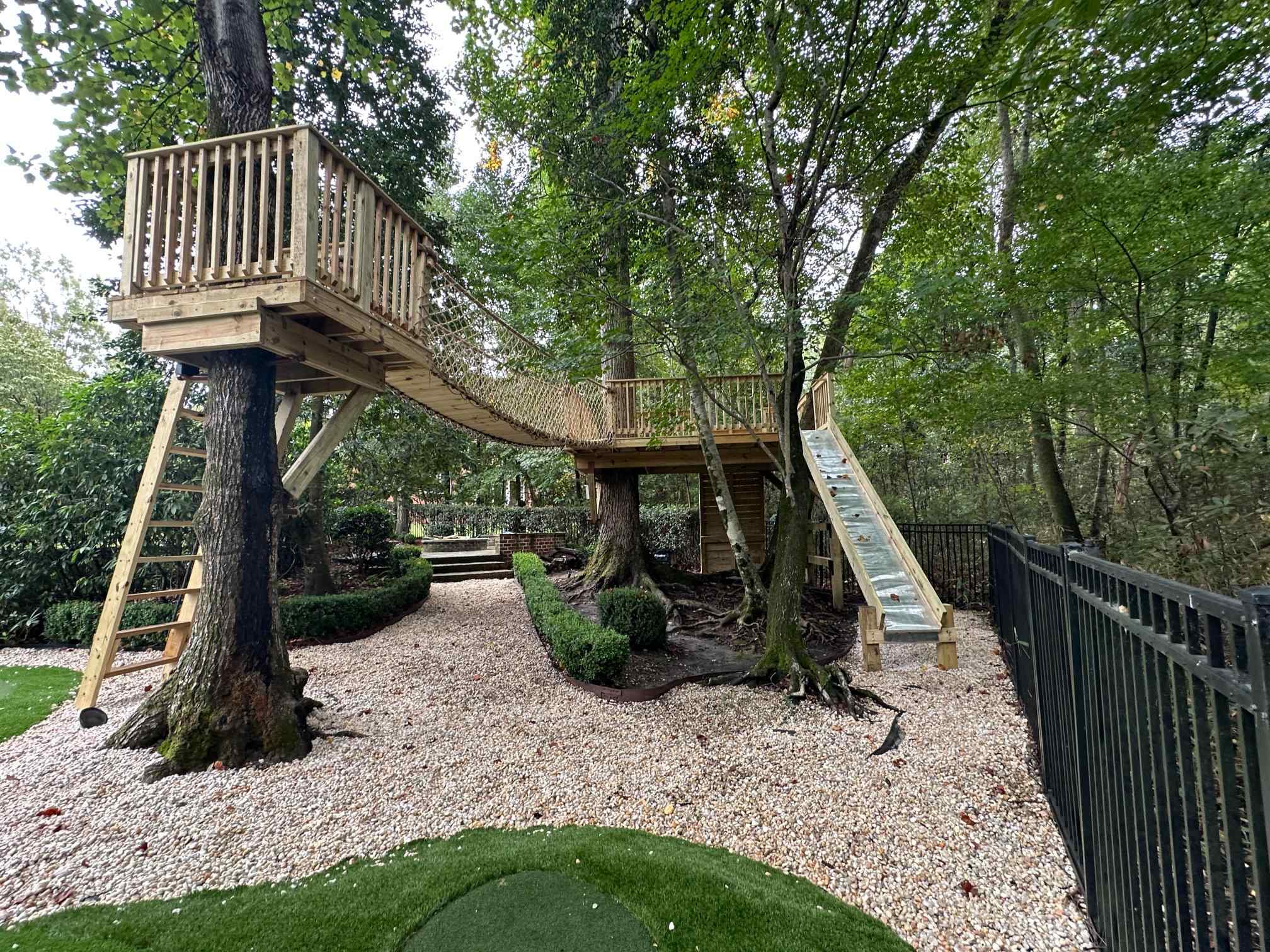 Two treehouse platforms built around mature trees, joined by a rope net suspension bridge, with a ladder and slide